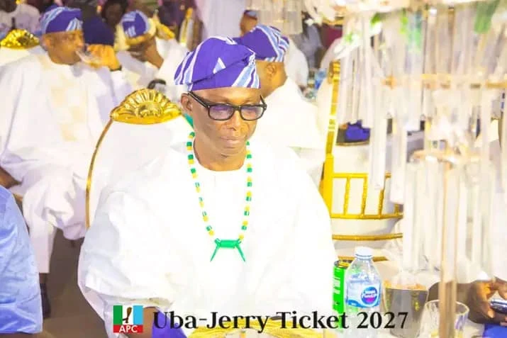 Jerry Adams seated in white attire, a purple cap and beads at a community gathering.