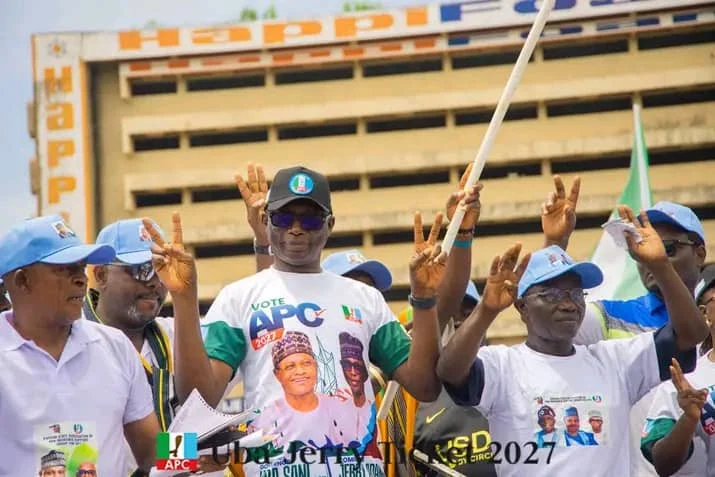 Jerry Adams raising his hands beside supporters in a Uba Sani–Jerry Adams campaign shirt.